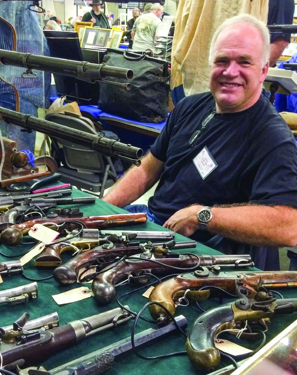 Al Duquette smiles for the camera. He sits behind a table full of antique firearms for sale.