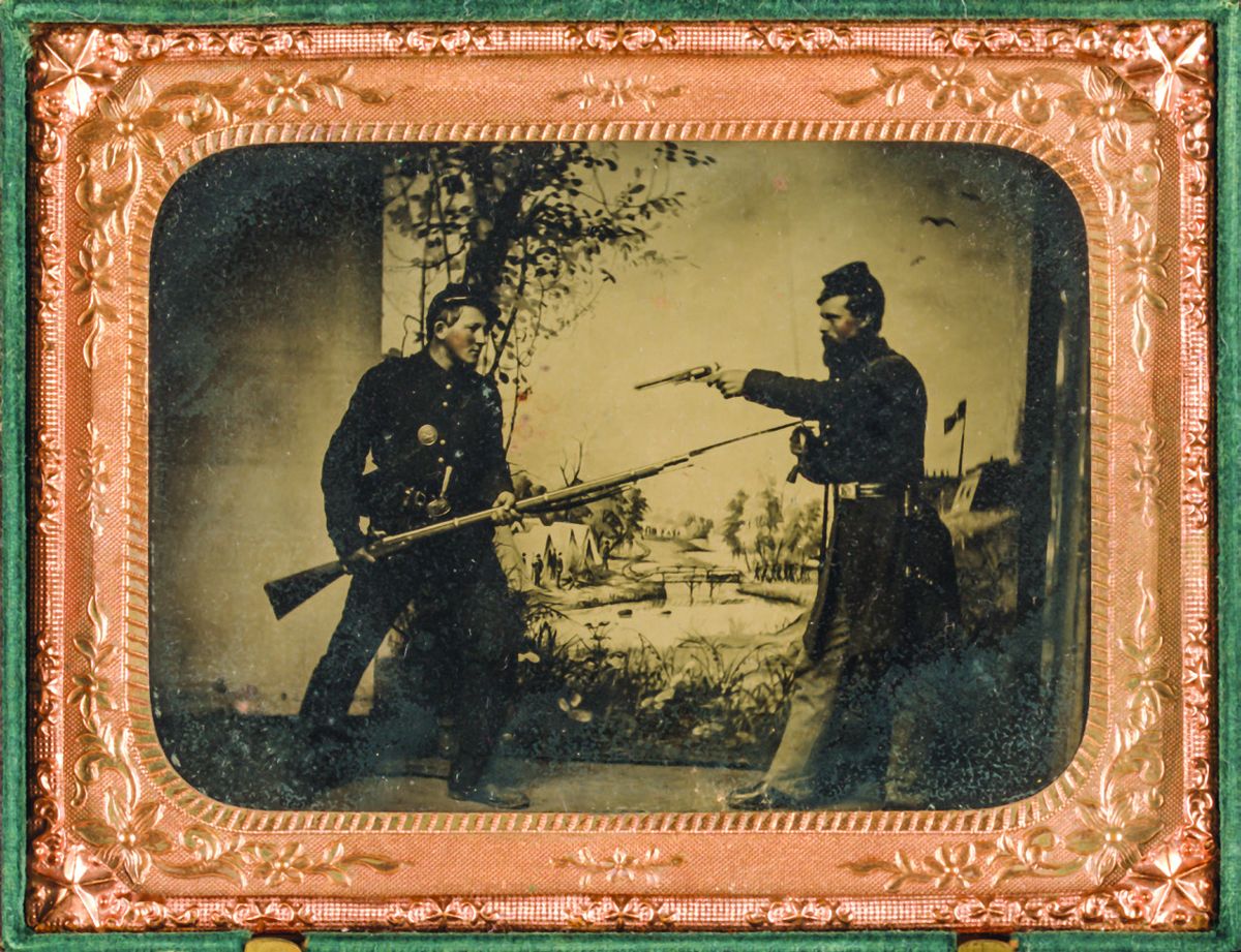 Two unidentified soldiers in Union uniforms posing with bayoneted Springfield M1861 rifled musket with attached bayonet, knife, and Colt M1851 Navy revolver in front of painted backdrop showing landscape with camp. (Library of Congress)