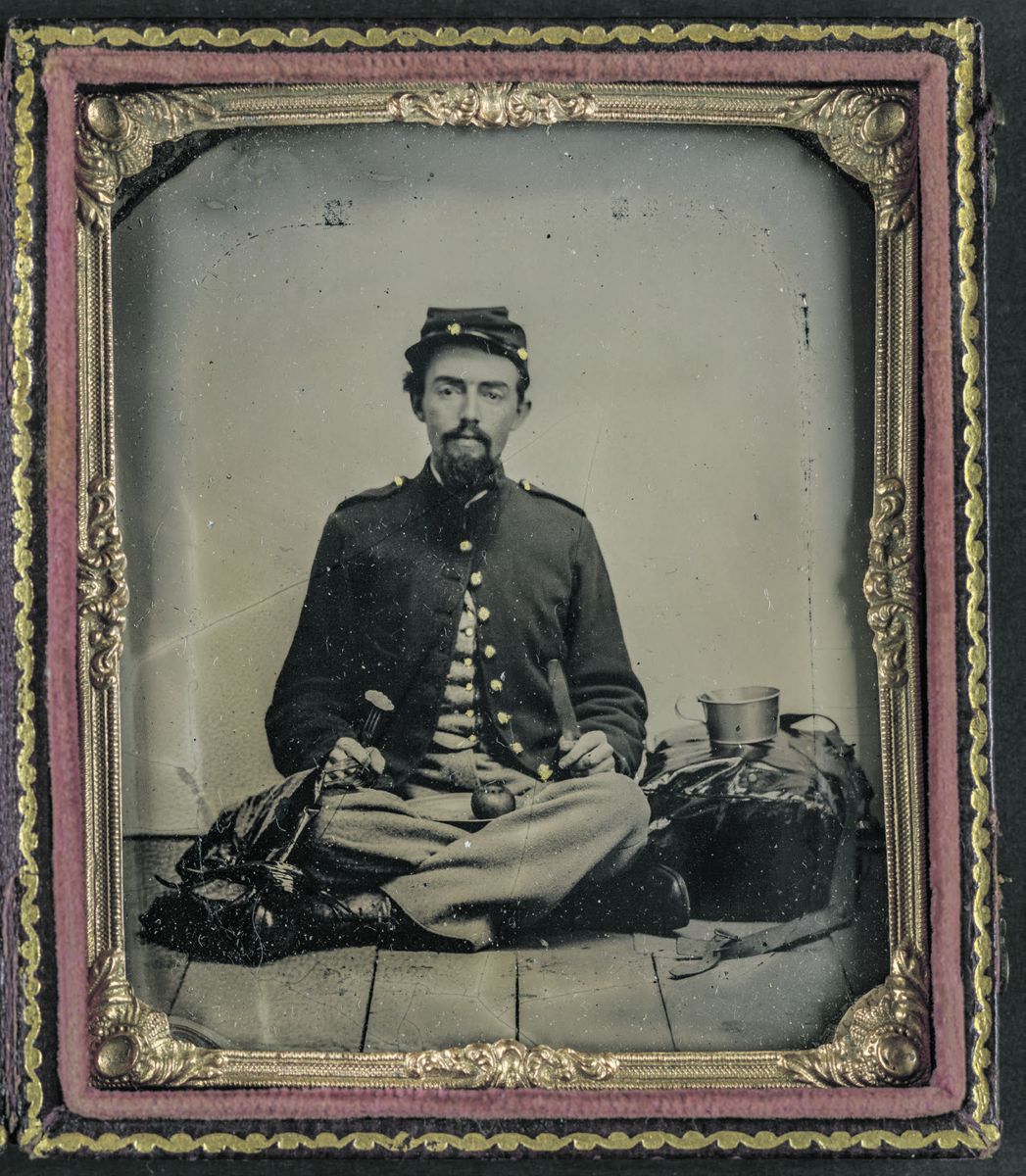 Unidentified soldier in Union uniform with fork, knife, plate, and cup sitting on the floor and preparing to eat a slice of the apple on his lap. Library of Congress. Liljenquist Family collection.