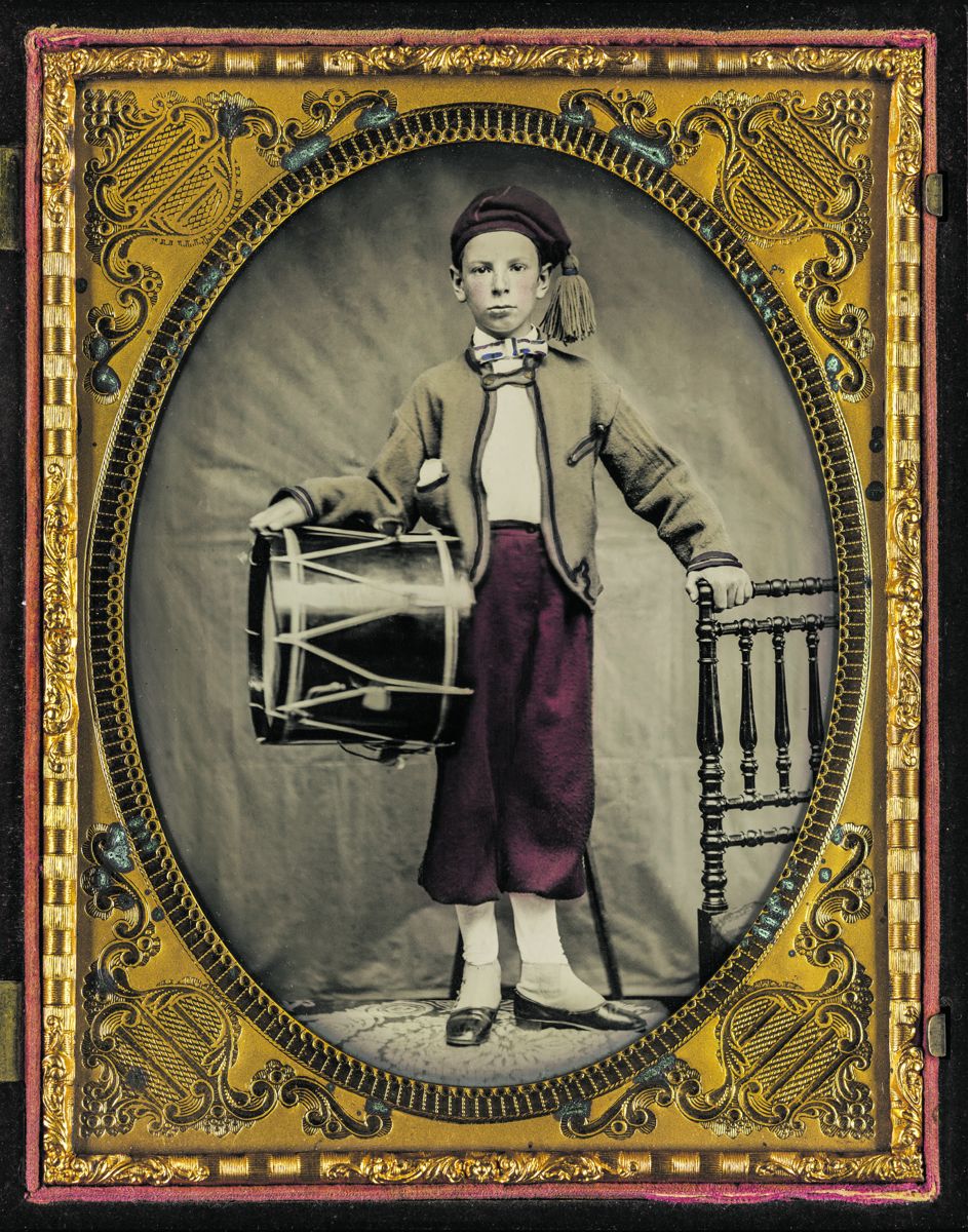 Unidentified boy wearing Zouave style uniform, holding a drum, and wearing a red white and blue bow tie. This young man might not be a drummer. His uniform is not fancy enough for a musician and most drummers held their drum sticks for a photograph. Note the pair of slit pockets on the jacket, and a handkerchief in the right side chest pocket. The lack of a belt or suspender lines indicates that the pantaloons are held at the waist by a tie or buckle mounted on the rear waistband, or connecting the two band ends above a gore.
