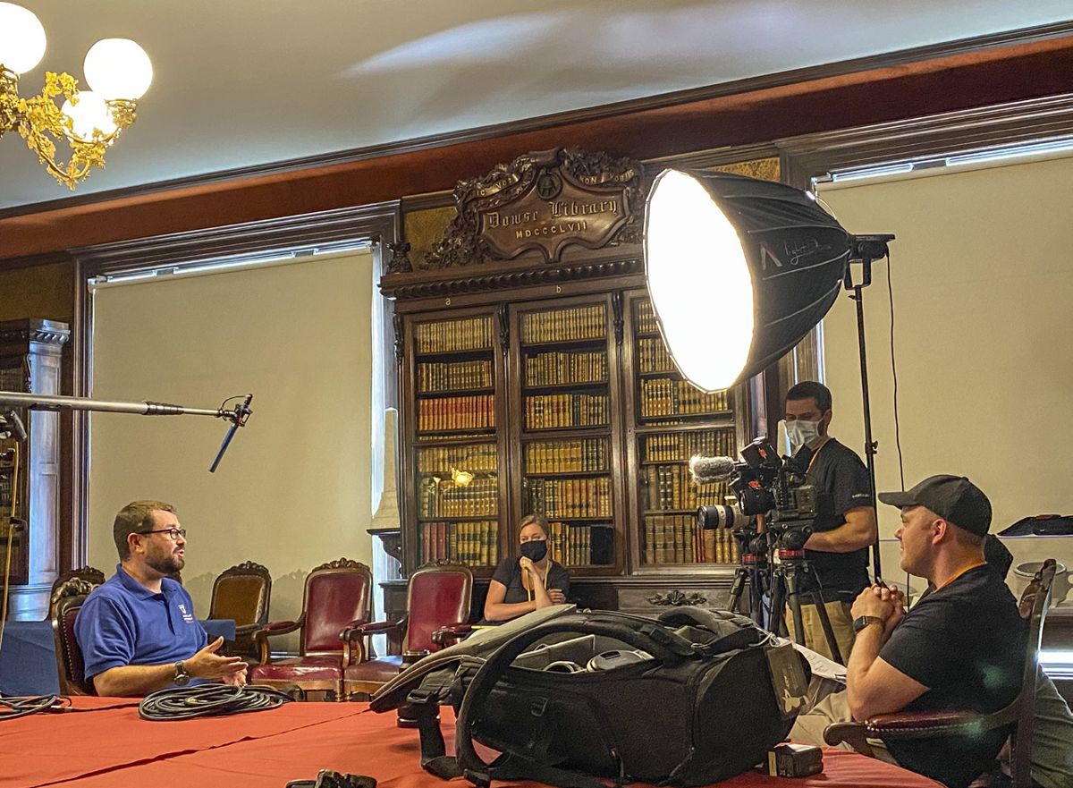Kris White, seated in a library at the Massachusetts Historical Society, talks with a film crew about Revolution-