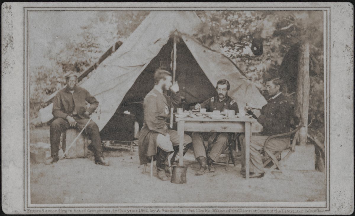 Three unidentified soldiers eating at table outside tent as an African American soldier sits by the tent with sword on his knee.